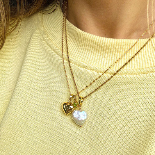 Pozłacane naszyjniki warstwowo na szyi modelki. Jeden z perłą w kształcie serca, drugi z serduszkiem i perydotem.
