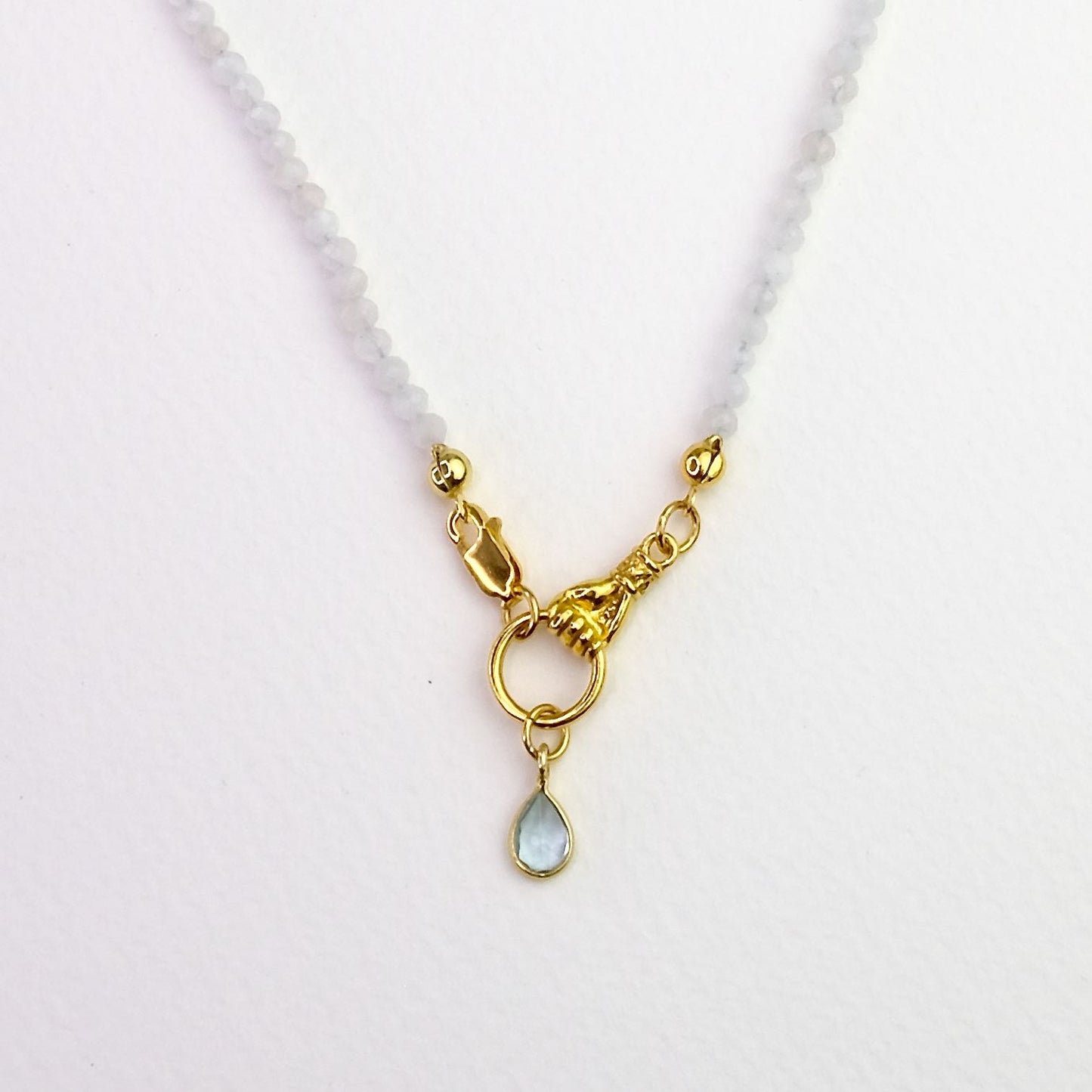 Naszyjnik z kamieni księżycowych ze złotą zawieszką w kształcie dłoni trzymającej obręcz na której zawieszony jest topaz, close-up na jasnym tle.